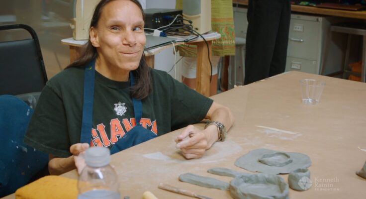 A Creativity Explored artist working with clay and smiling at the camera.