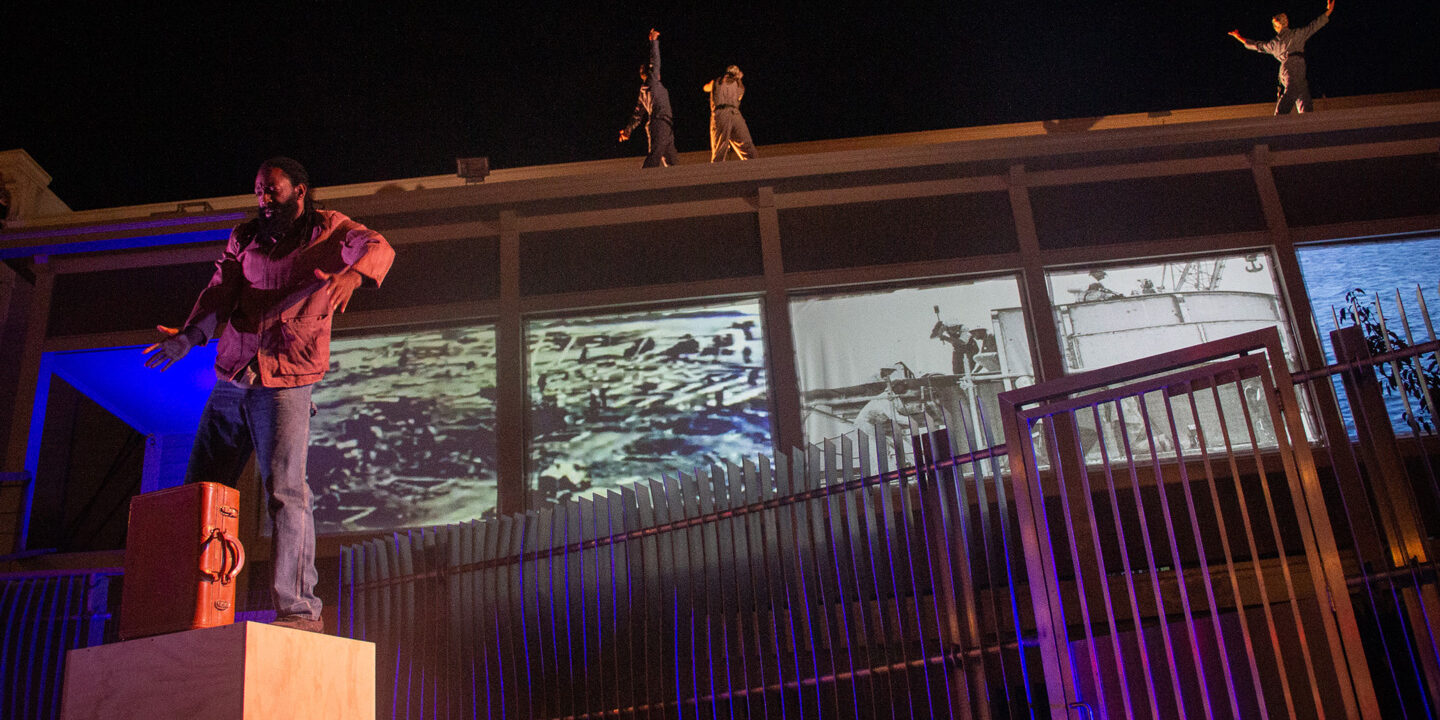 A site specific dance in Bayview-Hunter’s Point where three dancers are on top of a roof and one dancer is dancing on a wooden block with a briefcase.