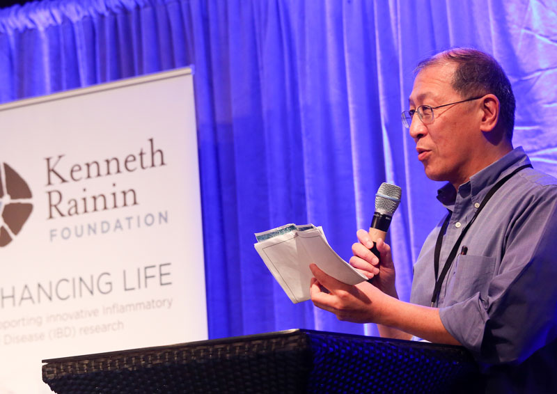 Person standing at a podium speaking into a microphone towards an audience. In the background a white banner with the Kenneth Rainin Foundation's logo hangs