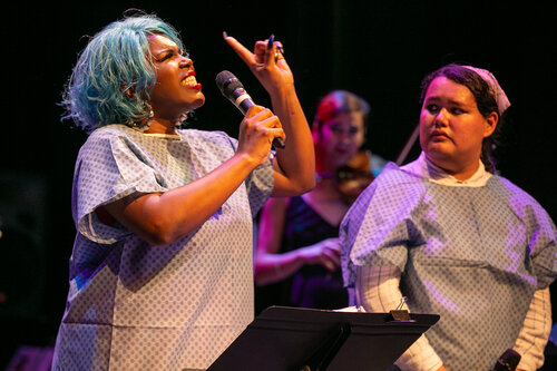 Two performers standing wear medical gowns, one speaks into a microphone