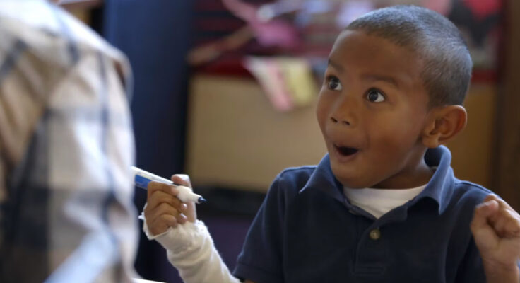 A young child raises both fists in the air with an excited expression on his face as he begins understanding how to write.