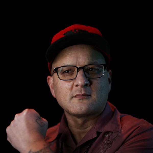 A male-appearing person with a raised fist, wearing a burgundy barong shirt, glasses, and a red and black United Farm Workers hat.