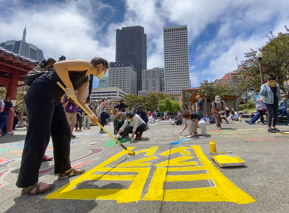 A woman wearing a mask, painting a large Chinese character in yellow on the pavement, in solidarity with Black Lives Matter.