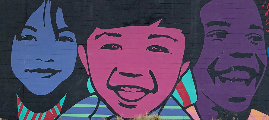 A closeup of the “We Are The Ones We’ve Been Waiting For” mural by Jessica Sabogal. It shows three children, smiling, and colored in shades of blue, pink and purple.
