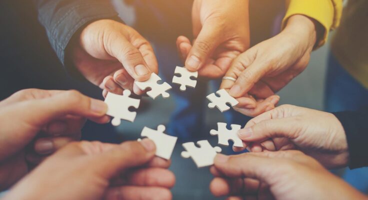 A circle of seven hands each holding a jigsaw puzzle piece, bringing them together in the center.