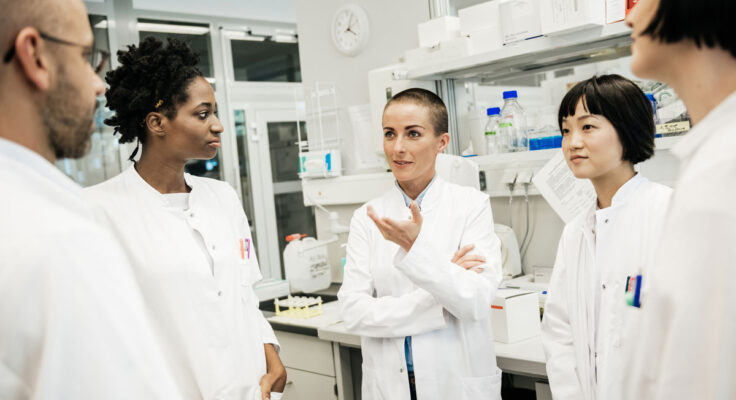 A diverse group of five researchers in white coats talking in their lab.