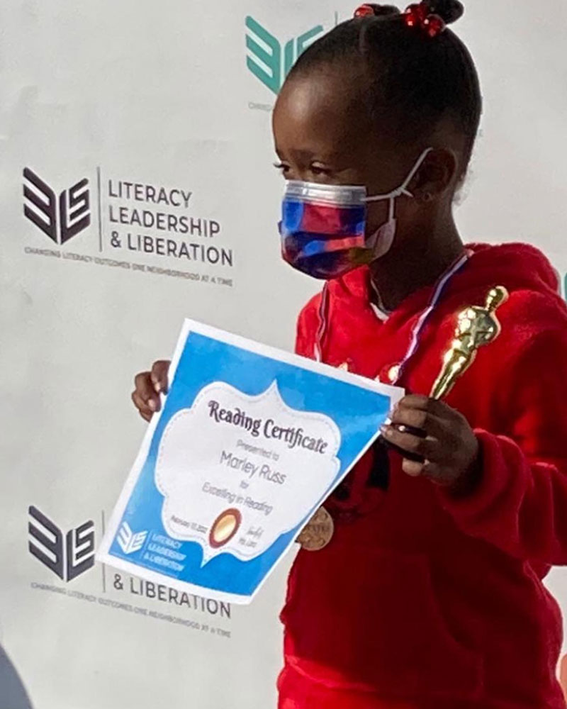 A young student wearing a red shirt and a medical mask proudly holds a paper reading certificate