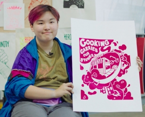 A person in a colorful jacket holds up a pink screen printing with the text, "cooking carries our stories."