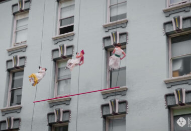 Three dancers wearing white dresses rappel down the face of a historic building