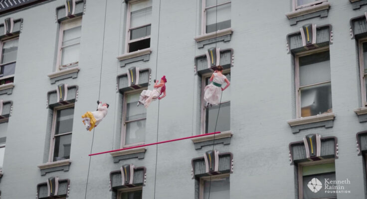 Three dancers wearing white dresses rappel down the face of a historic building
