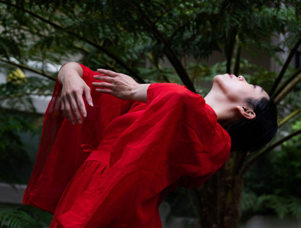 a dancer wearing a bright red gown leans backwards against a backdrop of lush greenery
