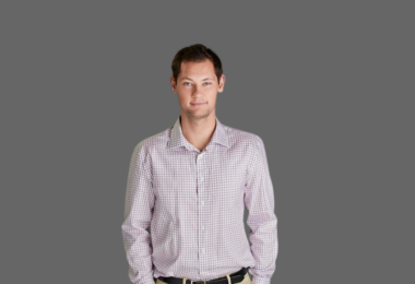 Person with very short, darker hair stands with hands in their pant pockets against a gray photo backdrop