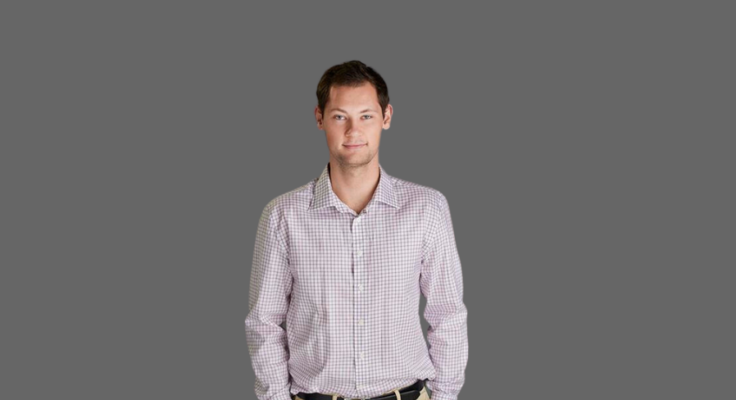 Person with very short, darker hair stands with hands in their pant pockets against a gray photo backdrop