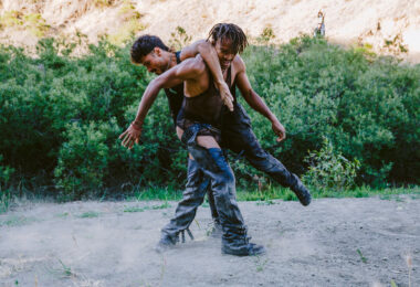 Two dancers stand connected on a patch of dirt. Both wear black leather pants and mesh tops. Their black boots kick up dust as they move on the ground.