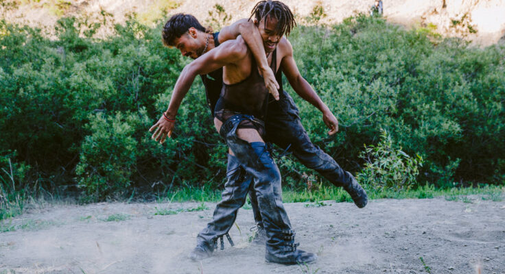 Two dancers stand connected on a patch of dirt. Both wear black leather pants and mesh tops. Their black boots kick up dust as they move on the ground.
