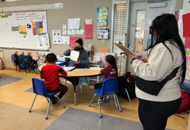 A parent watches as an educator works with two young children sitting at a shared desk.