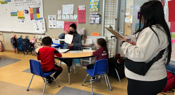 A parent watches as an educator works with two young children sitting at a shared desk.