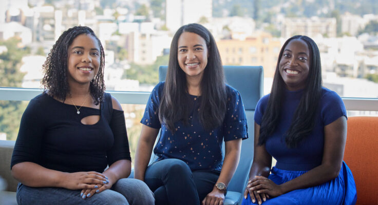 three people sit closely on a couch smiling and looking toward camera