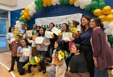 a group of 16 people, many holding paper certificates and smiling under an arc of balloons