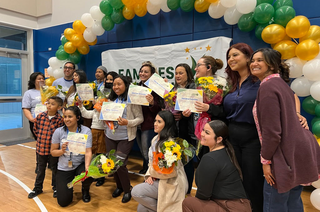 a group of 16 people, many holding paper certificates and smiling under an arc of balloons