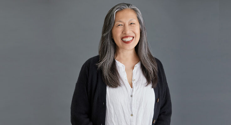 Against a gray background a person with chest length black and gray hair smiles toward the camera. She is wearing a black cardigan and white dress.