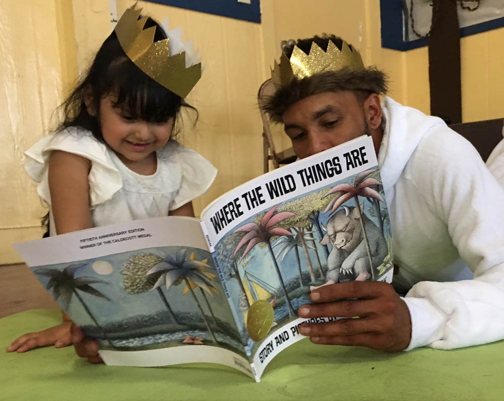 An an adult and a smiling child, both wearing gold paper crowns, lay and sit on the floor and read from "Where the Wild Things Are."