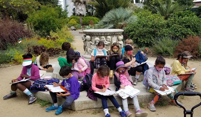 14 children sit, while writing on paper, in a park-like setting