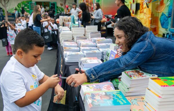 smiling adult leans over a long table filled with books to help a child. In the background young children stand with tote bags.