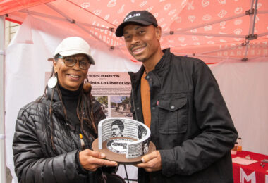 Two people smiling toward the camera and holding a three-dimensional c-shaped object printed with black and white photos and text on it.