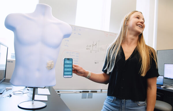 A person holding up their phone next to a wireless patch on a mannequin to demonstrate how the "EKG for the gut" works.