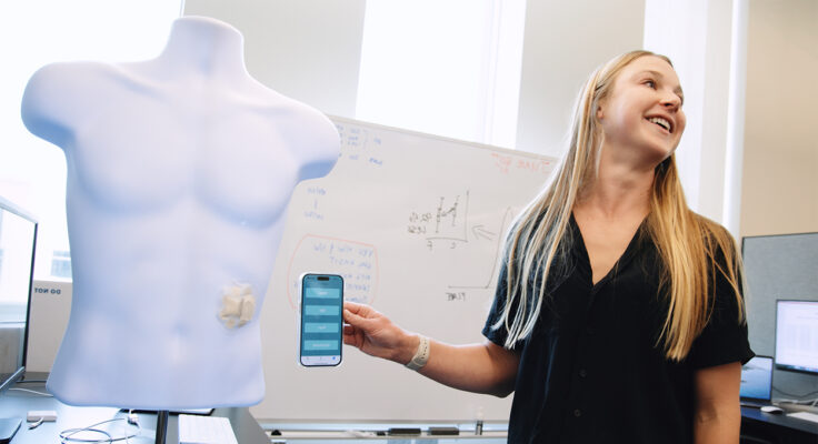 A person holding up their phone next to a wireless patch on a mannequin to demonstrate how the "EKG for the gut" works.