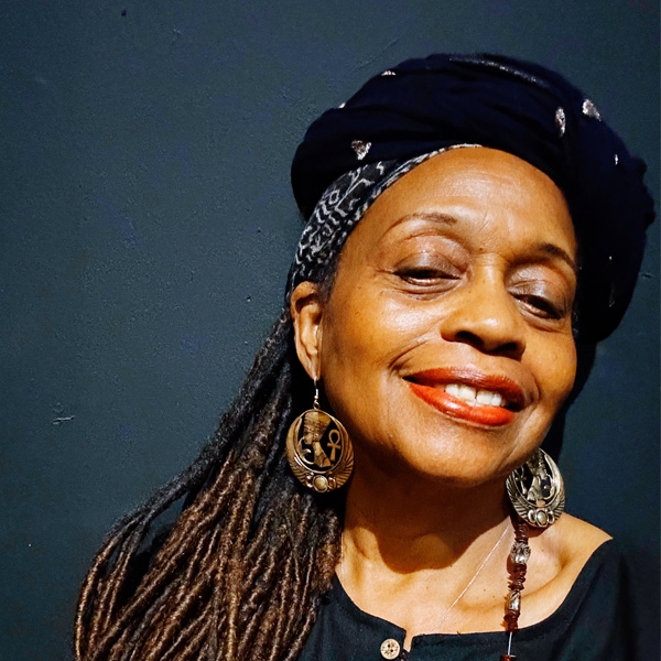 Ayodele, a smiling African American woman with shoulder-length locs, wears a black blouse with wooden buttons on the front, a black and white patterned headscarf, and round, dangling earrings.