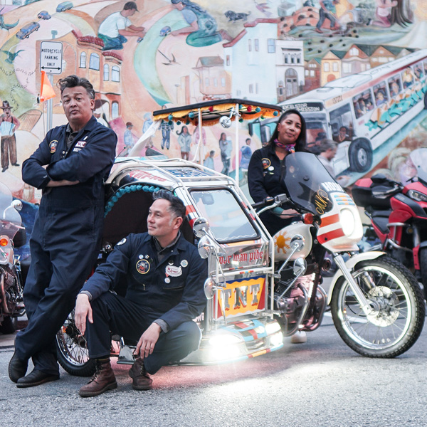 Two men and a woman wearing mechanic jumpsuits stand in front of a mural and pose around a traysikel, or Filipino motorcycle and sidecar. Behind them, several other motorcycles are parked in front of the mural.