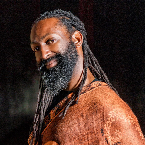Antoine, a Black and Indigenous person with almond-shaped eyes, long lashes, dark brown skin, a full beard, and long black locs tied in a low braid, wears a brown shawl and smiles.