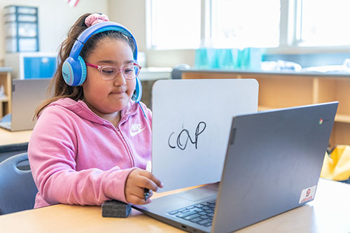 A student holds a whiteboard with the word "cap" written on it up to a laptop camera.