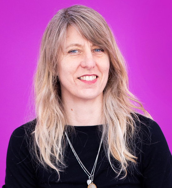 A white woman with long blond hair and bangs smiles against a neon purple background.