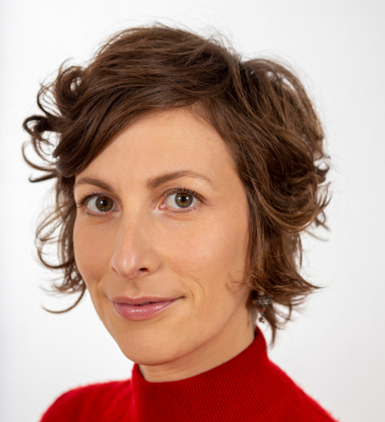 A woman with short brown hair, green eyes, and olive skin wears a red top.