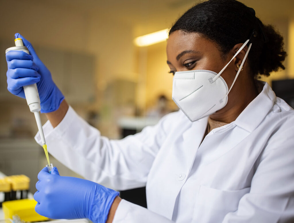 person in a white lab coat and medical mask uses a pipette