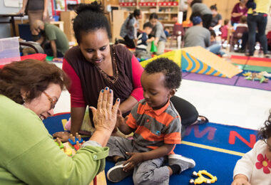 An early education classroom where two adults are on a playmat with two young children. One adult is high-fiving one of the children.