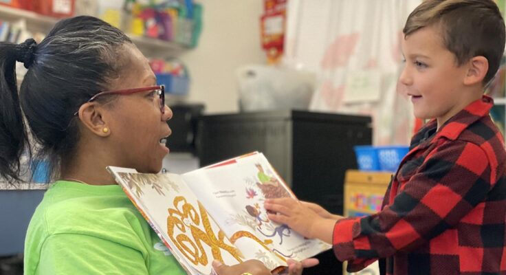 a smiling child points at a book that their teacher is holding open.