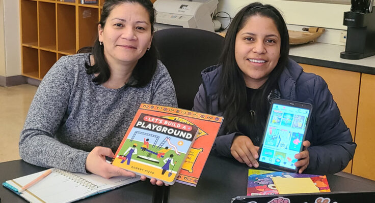 Two people are seated and smiling facing the camera holding children's books and a tablet