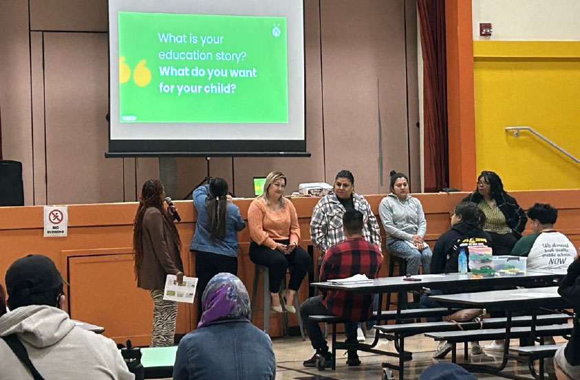 A panel of six people sit below a projection that reads "What is your education story? What do you want for your child?" The audience sits at long tables facing them.