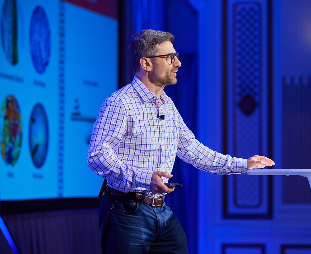 Person presenting in front of large projected image. The audience is not in the photo.