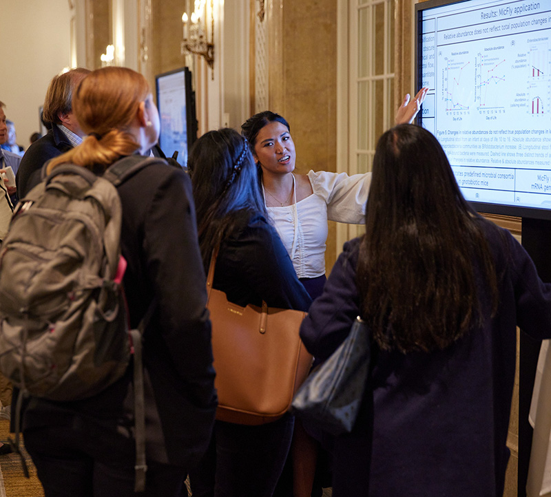 A person gestures toward a wall monitor showing scientific data while four people listen intently. In the background you see more monitors and people.