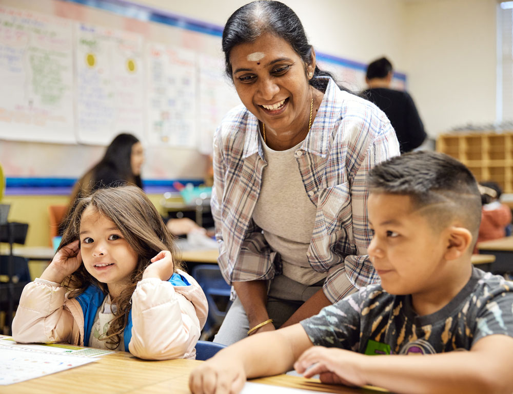 A smiling adult crouches near two young children. One of the children smiles at the camera.