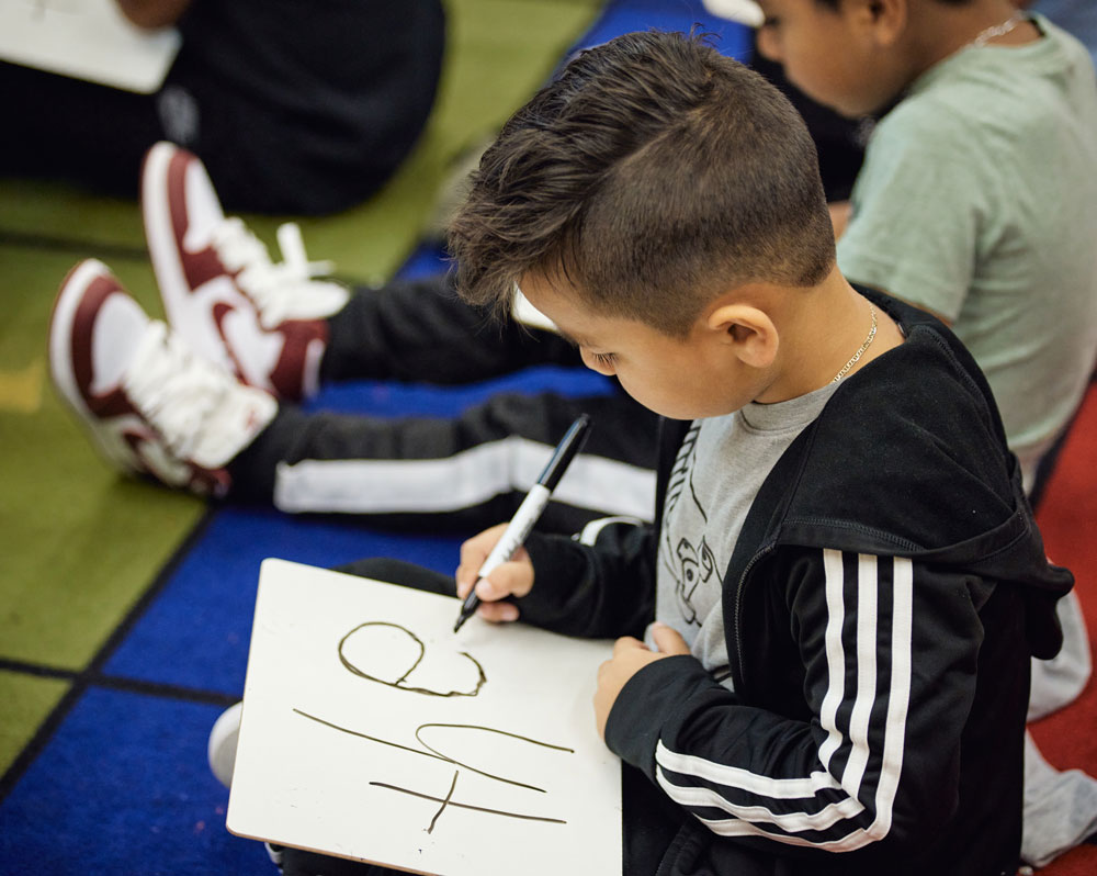 A young student sits on colorful carpeting, practicing writing the word 'the' on a small whiteboard.