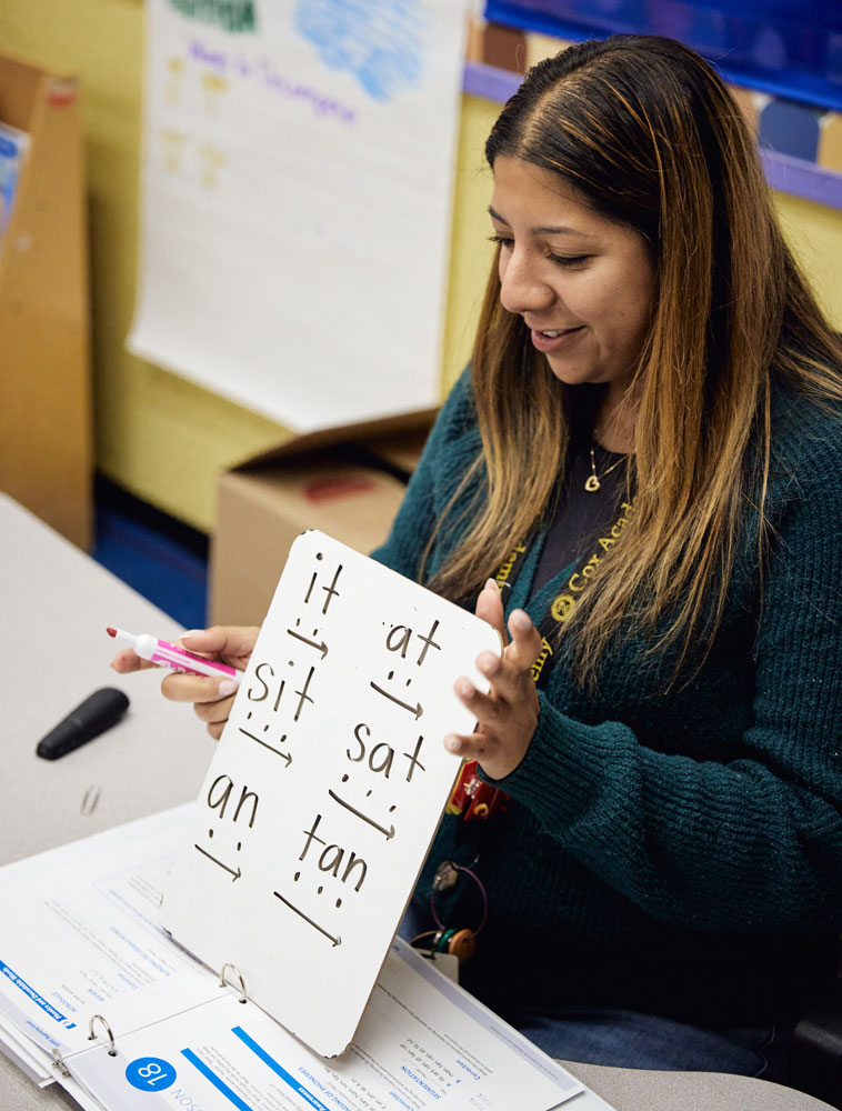 An adult uses a small white board to explain and compare sounds: it to at, sit to sat, and an to tan.