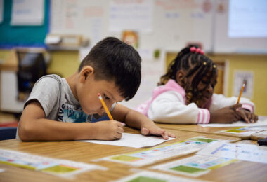 Two young students lean over their desks, actively focused on writing