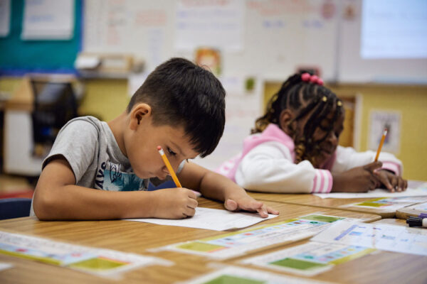 Two young students lean over their desks, actively focused on writing