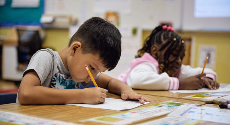 Two young students lean over their desks, actively focused on writing